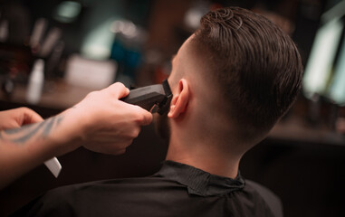 A barber uses clippers to trim a client's beard and neckline in a modern barbershop, showcasing...