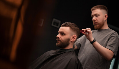 A young barber styles a male client's freshly cut hair with a comb in a modern barbershop, both focused and calm in a well-lit, professional grooming environment..