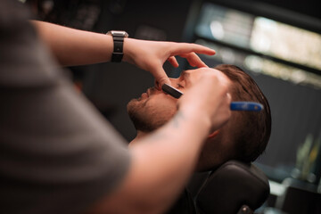 A barber precisely shapes a client's mustache with a straight razor, using one hand to stretch the skin for accuracy. The client sits calmly with eyes closed during the grooming session..