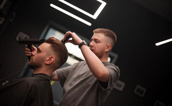 Young male barber styling a client's hair using a hairdryer and brush in a modern barbershop with geometric ceiling lights and a dark interior design. Professional grooming in progress.
