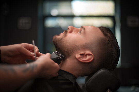 A young man reclines in a barber chair while receiving a precise beard trim with electric clippers in a modern barbershop. The focus is on grooming, style, and professional care..