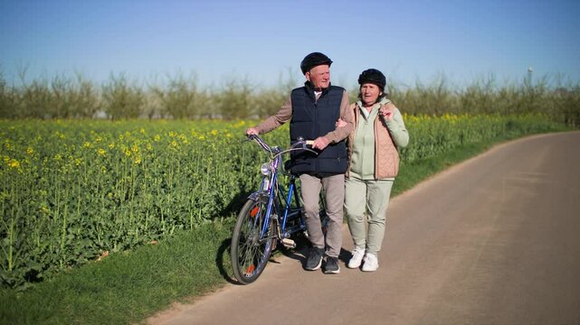 An elderly couple dismount from bicycle walks along a road in the countryside. 