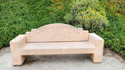 Beige colored concrete park bench with a curved backrest set against a backdrop of green Ice Plant and Bladderpod near cliff slope, San Diego, California