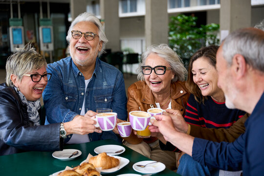 Group laughing mature people having snack sitting at cafe bar. Older friends toasting with coffee cups gathered together in terrace restaurant. Food and beverage lifestyle in senior citizens concept
