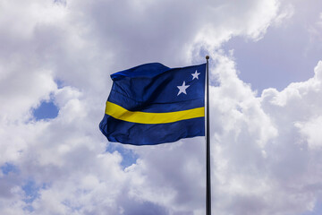 Curacao national flag waving on tall flagpole against cloudy sky with sunlight breaking through clouds. Willemstad. Curacao.