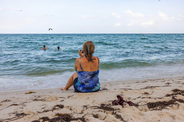 Woman sitting on sandy beach taking photo of swimmers in Atlantic Ocean in Miami Beach. 
