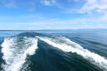 Wide view of boat wake trail on deep blue lake with distant mountain range and scenic summer sky, symbolizing travel, movement and freedom on the water