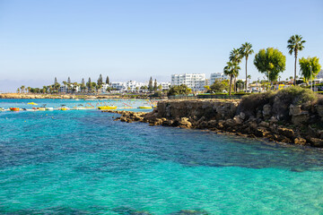 Beautiful coastal view of a busy beach with turquoise water, rocky shore, and white buildings in Cyprus...