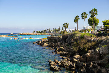 Beautiful coastal view of a busy beach with turquoise water, rocky shore, and white buildings in Cyprus.
