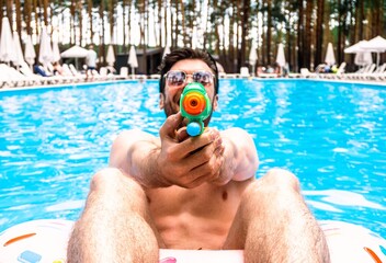 Happy guy playing with toy water gun while spending time in the swimming pool outdoors. Summertime, holidays, lifestyle concept