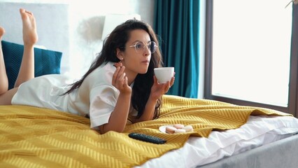 Beautiful Caucasian brunette woman in glasses eating macarons and drinking coffee or tea resting on comfortable bed watching TV. Slow motion. Food concept. Young female eats desert cookies in bed