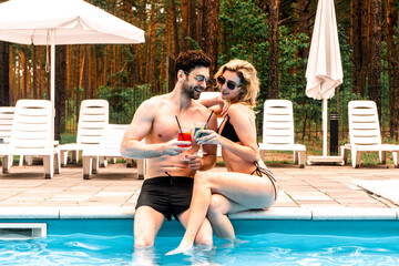 Smiling woman and her pleased boyfriend clinking glasses with cocktails while sitting on the swimming pool edge. Rest and relaxation concept
