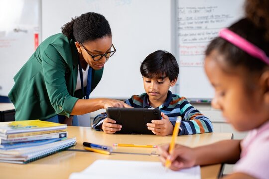 Teacher assisting young boy with tablet in classroom while girl writes with pencil in foreground, focused and engaged learning environment