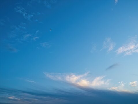 Clear blue sky with wispy white clouds and a small crescent moon during daylight
