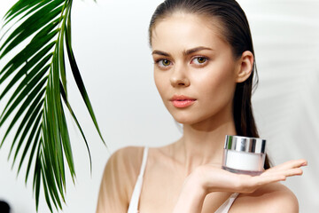 Young woman holding moisturizer with tropical palm leaves, showcasing skincare in a bright, fresh setting with natural beauty.