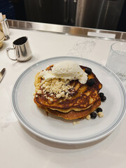 Gourmet blueberry pancakes with crumble topping and whipped cream, stacked breakfast pancakes served on white plate at brunch counter, luxury cafe brunch, sweet indulgent breakfast dish