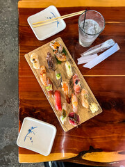 Top-down view of assorted nigiri sushi platter on wooden board with chopsticks, soy sauce dishes, and iced drink in Japanese restaurant setting