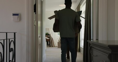 After moving in Diverse couple carrying box down hallway, unpacking pillows, throw, teddy bear