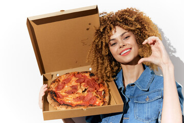 Cheerful young woman with curly hair joyfully holding a delicious pepperoni pizza in an open box, dressed in denim, against a bright white background