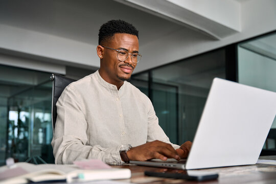 Busy smiling professional African business man company employee, young male worker businessman wearing glasses typing on computer using laptop working at office, elearning at workplace desk.