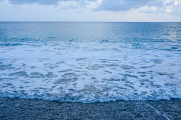 Obraz premium Turbulent Waves Meeting a Dark Pebble Shoreline: A dynamic scene of foamy ocean waves reaching a beach composed of dark pebbles. Coastal Scene: dark pebbles and foaming ocean waves.