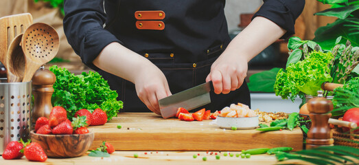 Chef cooking food in the kitchen. Cook cuts fresh strawberries, prepare salad. Wooden table,...