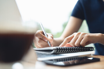 Business woman sitting at table in cafe, writing in notebook with laptop computer, mobile phone on table. Freelancer working in coffee shop. Student learning online, doing research and taking note