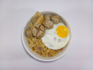 A white bowl filled with instant noodles, garnished with a fried egg, various meatballs, and some...