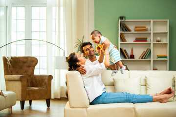Parents catch baby boy while sitting on sofa at home, enjoying fun playful family bonding time