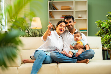 Fototapeta premium Happy Indian parents with newborn baby pointing sideways while relaxing on modern indoor sofa