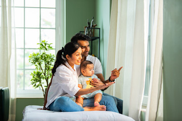 Mother pointing outside while sitting on bed with baby boy and father near sunny home window