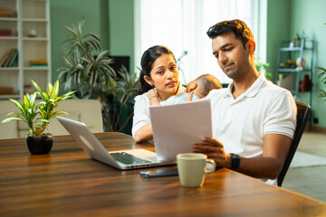 Indian father works on laptop as wife holds baby beside him, couple handling home responsibilities