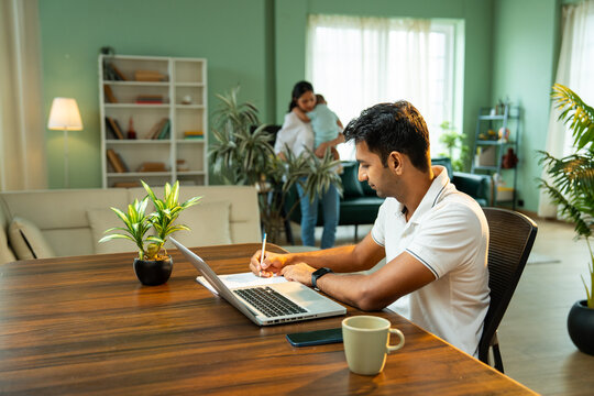 Father working on laptop at home while mother walks holding newborn baby boy indoors