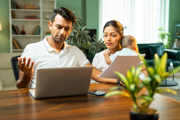 Stressed Indian father working on laptop while mother walks with newborn baby boy indoors at home