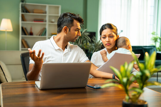Stressed Indian father working on laptop while mother walks with newborn baby boy indoors at home