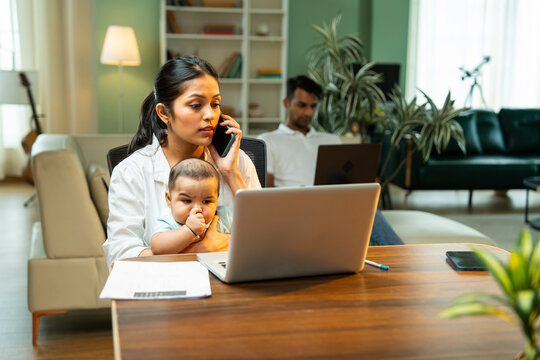 Mother working on laptop holding baby boy while father walks in background indoors at home