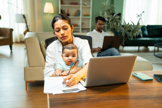 Mother working on laptop holding baby boy while father walks in background indoors at home - Powered by Adobe