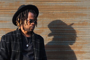 Modern adult black man with dreadlocks wearing hat and stylish coat against street wall in sunlight