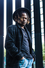 Cool African American male with dreadlocks and in hat holding hands in pockets of denim looking at...