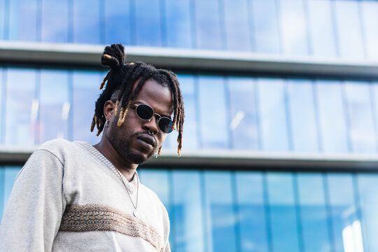 Modern African American man in sunglasses and dreadlocks in bun hairstyle looking at camera against skyscraper - Powered by Adobe