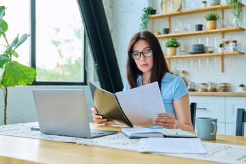 Young serious woman working with laptop, reading business papers, sitting at table at home
