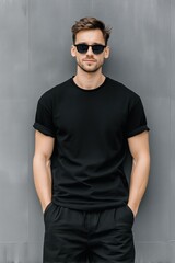 Naklejka premium A stylish young man wearing a black t-shirt mockup and sunglasses, standing confidently against a gray wall. His casual look and relaxed posture convey a modern, urban vibe.