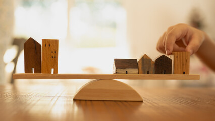 A child's hand delicately places a house model on a balance, weighing it against a pile of money,...