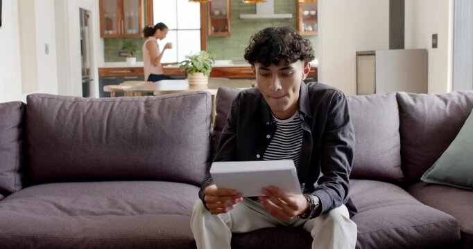Diverse mother and son opening envelope and unfolding letter on gray sofa sharing exciting news