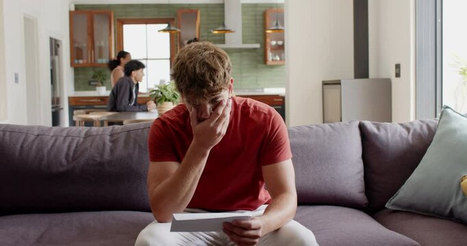 White man grabbing letter from sofa in kitchen reading letter anxiously while friends gathering