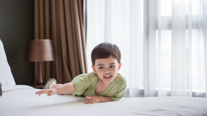 cheerful young boy in a green shirt smiling while lying on a bed in a cozy bedroom. Natural light...