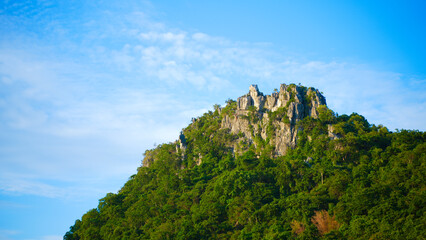 Beautiful rocky mountain covered in lush green trees under a bright blue sky with light clouds. A natural landscape perfect for travel themes, eco-tourism, environment conservation, and outdoor advent