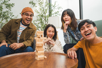 Young Asian friends are happily playing wooden block together at home, enjoying their leisure time. Group friends carefully remove a wooden block while others watch, having fun a classic table game.