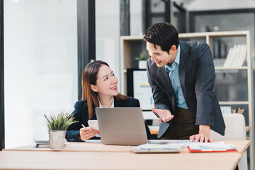 Two business colleagues smiling and discussing work together in modern office with laptop and documents on desk, showing positive and collaborative atmosphere
