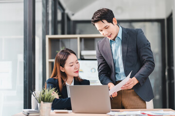 Two business colleagues discussing documents in modern office with laptop and charts, showing collaboration and professional teamwork in bright workspace